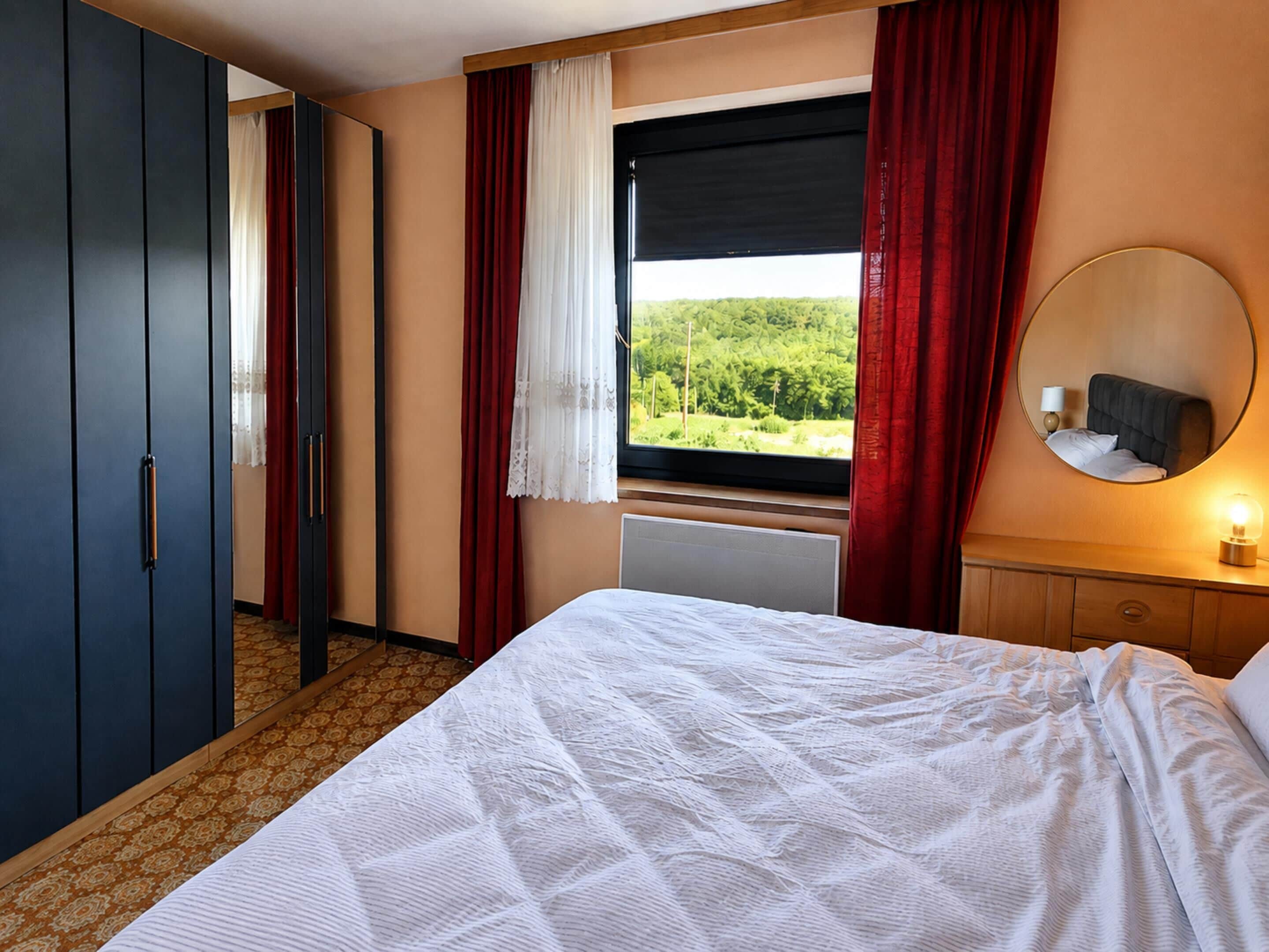 A hotel room with a bed, a window with a view of greenery, a wardrobe, and a bedside table with a lamp.