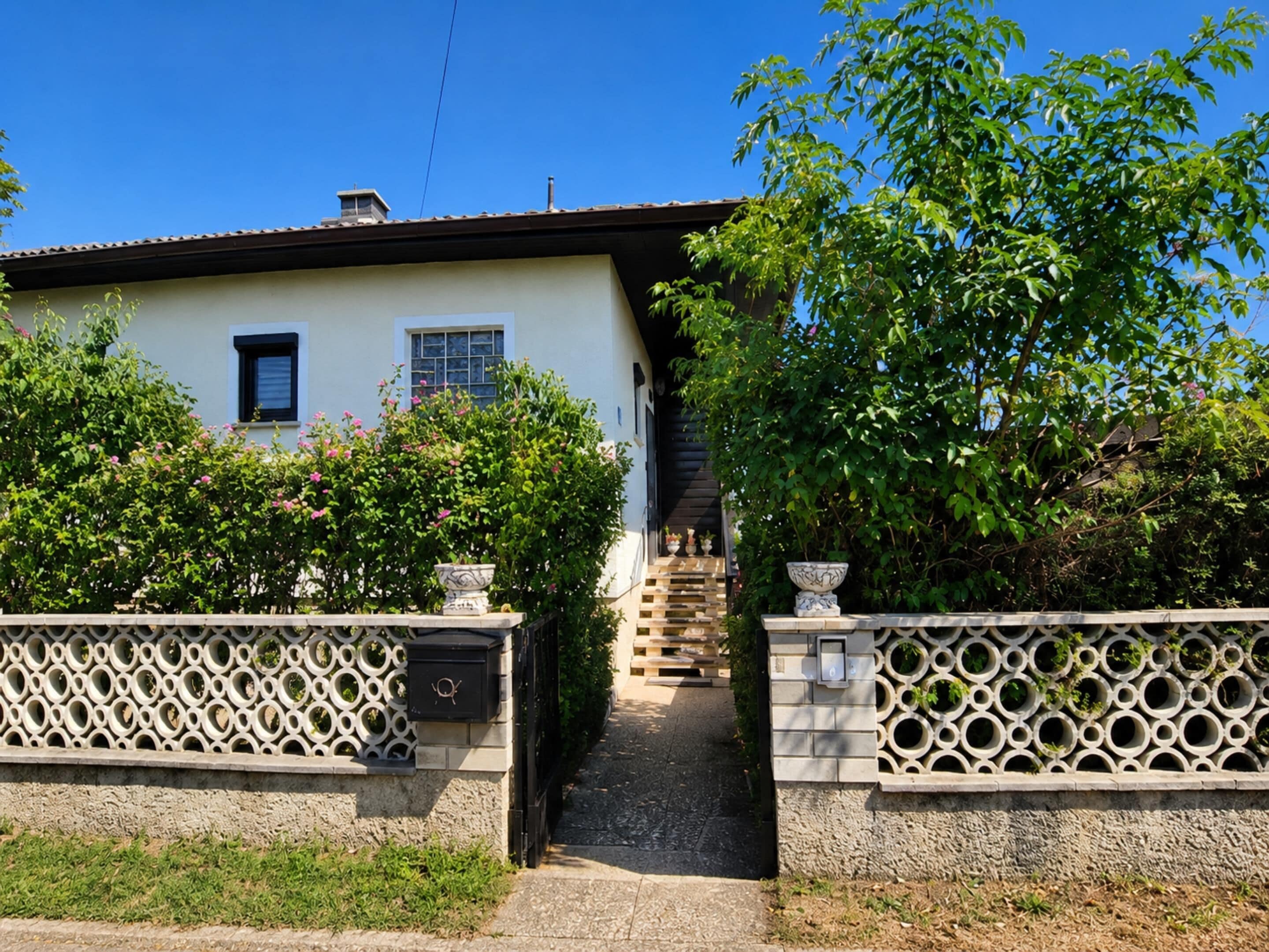A house with a paved entrance, a staircase leading to the door, and a garden with a fence.
