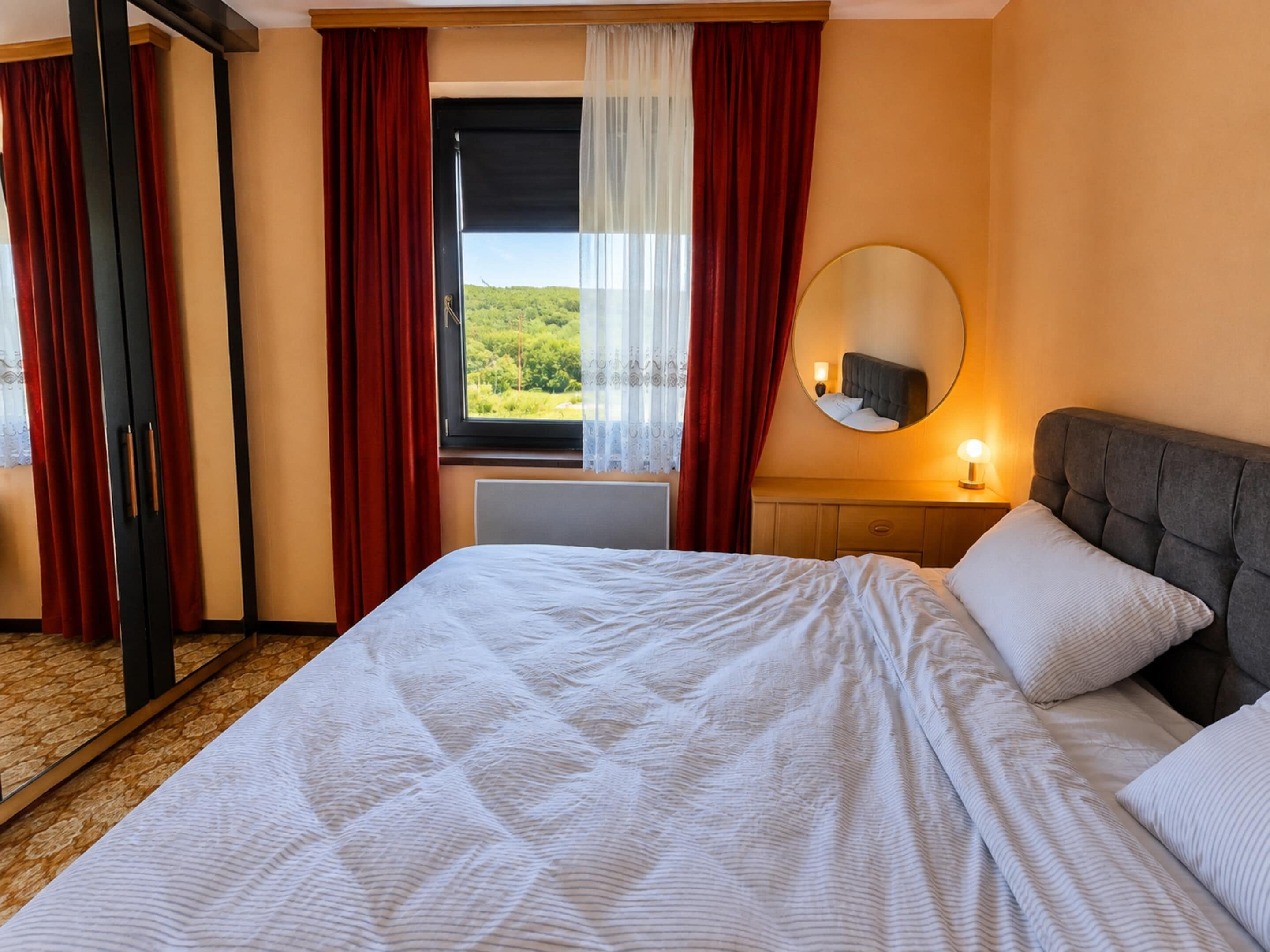 A hotel room with a bed, a window with curtains, a bedside lamp, and a mirror.