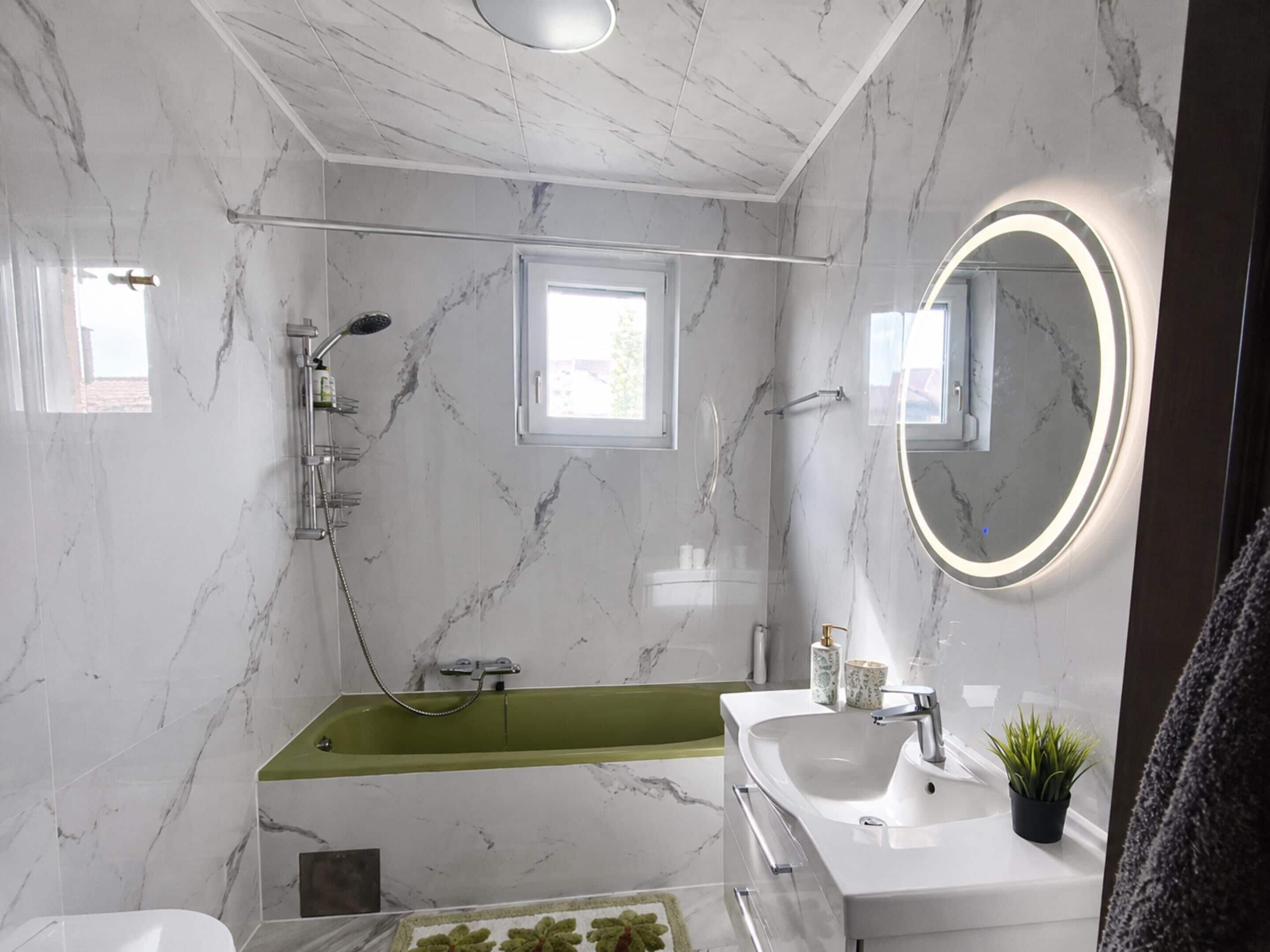 A modern bathroom with a marble wall, a green bathtub, a white sink, and a round mirror with a light.