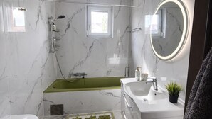 Ein modernes Badezimmer mit Marmorwand, einer grünen Badewanne, einem weißen Waschbecken und einem runden Spiegel mit Beleuchtung.