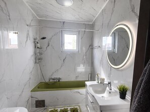 Un bagno moderno con una parete in marmo, una vasca da bagno verde, un lavandino bianco e uno specchio rotondo con illuminazione.