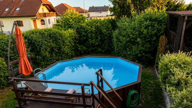 Ein sechseckiger Pool mit Holzterrasse und Liegestühlen, umgeben von Grünflächen und Wohnhäusern.