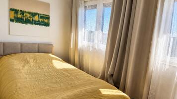 Ein Schlafzimmer mit Bett, Kopfteil, einem Fenster mit Vorhängen und einem Gemälde an der Wand.