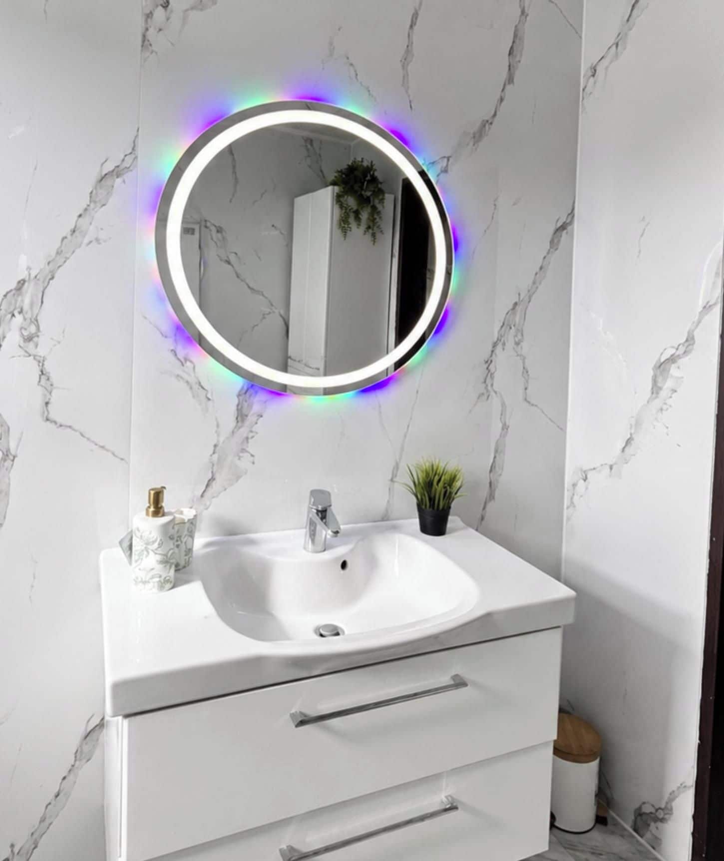 Une salle de bain avec un lavabo en marbre blanc, un miroir doté d’un éclairage LED circulaire et une petite plante en pot.