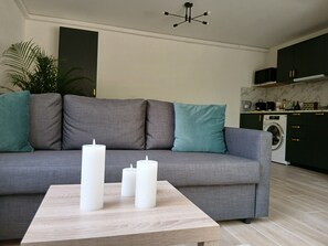 A modern living room with a grey sofa, teal cushions, a wooden coffee table, and three white candles. - Rose Stone: 2-Bedroom Apartment with Pool, Where Land Meets Sea on the Riviera (Opio)