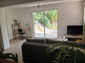 Ein Wohnzimmer mit einer Couch, einem Schreibtisch mit Stuhl, einem Fernseher und einer Schiebetür zu einem Außenbereich mit Pflanzen.