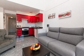 A modern living room with a grey sofa, a coffee table with fruit, and a kitchen with red cabinets. - Central 1 Bedroom Apartment. Communal Heated Pool (Los Cristianos)