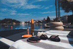 Une chaise longue sur une plage, avec une bouteille, des lunettes de soleil et un téléphone, offrant une vue sur la mer et les bâtiments.