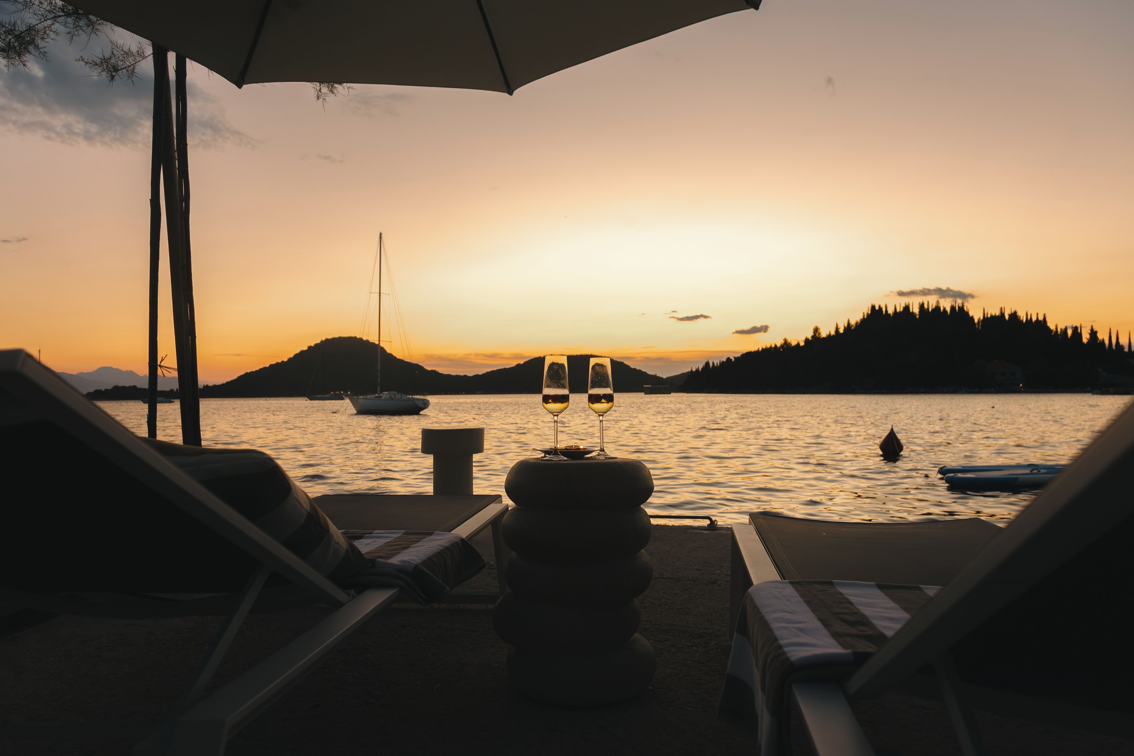 Une vue sur le coucher de soleil depuis un salon de plage, avec deux verres de vin sur une table, donnant sur des eaux calmes et des bateaux.
