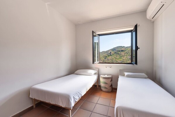 A small, clean room with two single beds, a window with a view of a mountainous landscape, and an air conditioning unit.