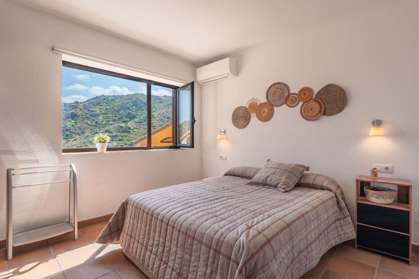 A bedroom with a large window, a bed with a plaid duvet, a wall-mounted shelf, and a wall-mounted lamp.