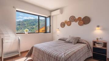 A bedroom with a large window, a bed with a plaid duvet, a wall-mounted shelf, and a wall-mounted lamp.
