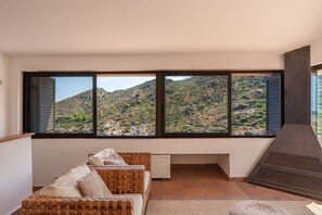 A room with a large window overlooking a mountainous landscape, a wicker sofa, and a fireplace.