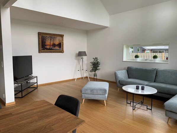 A modern living room with a flat-screen TV, grey sofa, round coffee table, and a dining area with a wooden table.