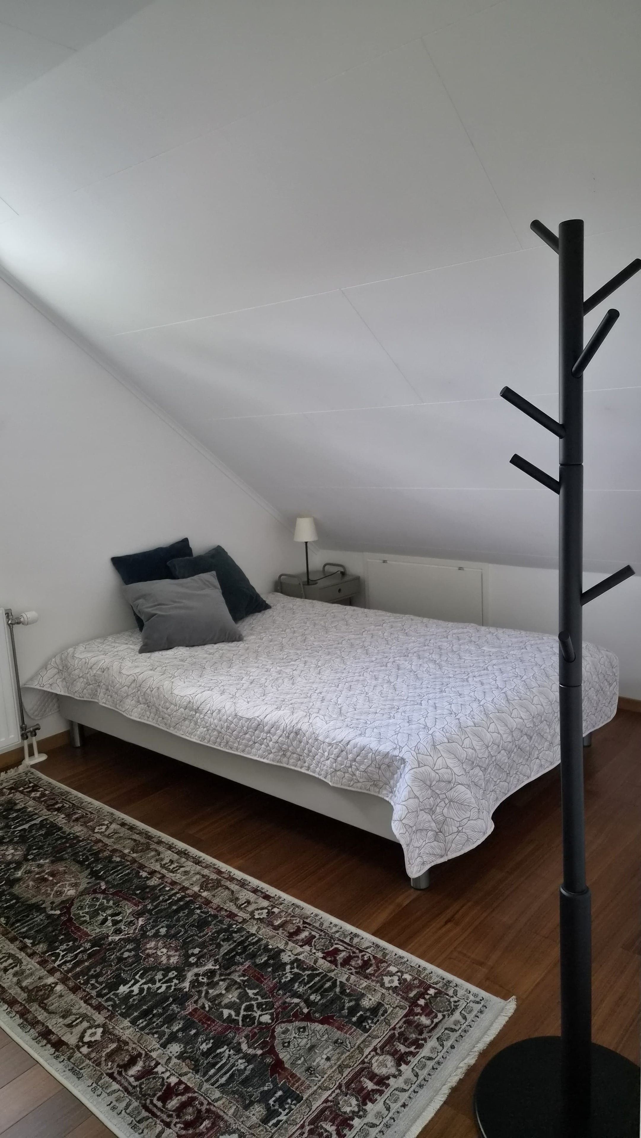 A bedroom with a bed, a nightstand, a lamp, a coat rack, and a patterned rug.