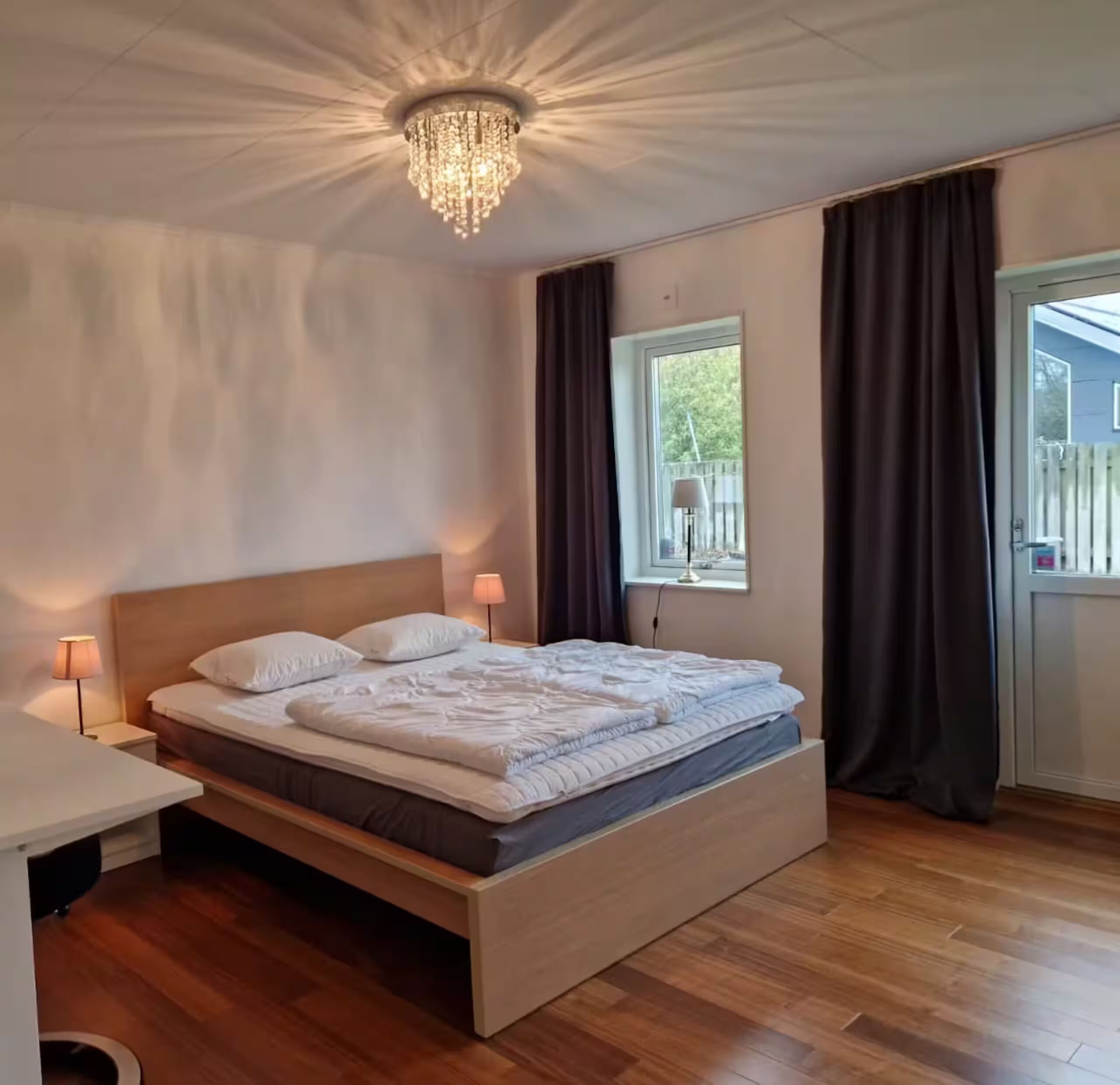 Una camera da letto con un letto di legno, un comodino, una lampada, un lampadario, una finestra con tende e vista sull'esterno.