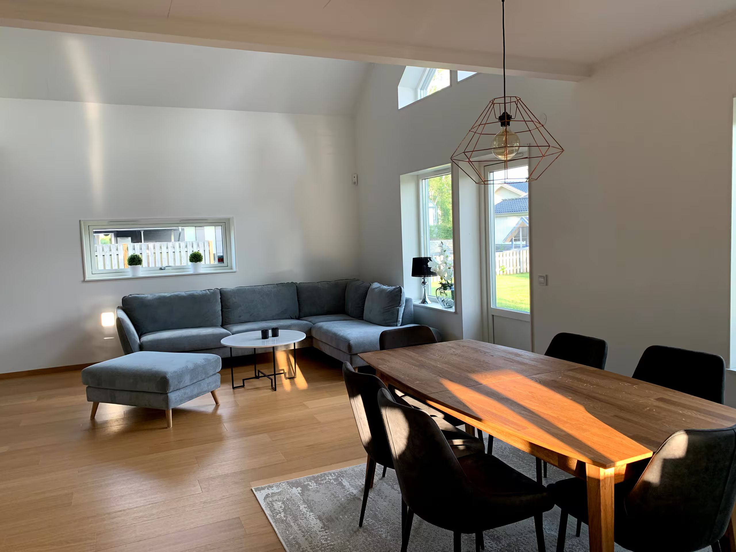 Ein zeitgemäßer Wohnbereich mit einem Ecksofa, einem runden Couchtisch, einem Esstisch mit Stühlen und einer Pendelleuchte.