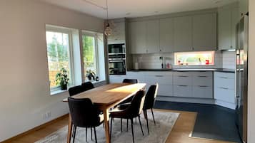 Eine moderne Küche mit weißen Schränken, eine Essecke mit einem Holztisch und Stühlen sowie ein Blick ins Freie durch große Fenster.