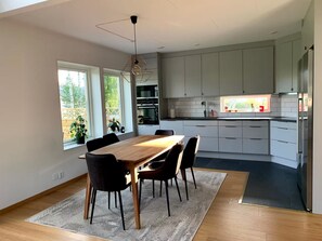 Eine moderne Küche mit weißen Schränken, eine Essecke mit einem Holztisch und Stühlen sowie ein Blick ins Freie durch große Fenster.