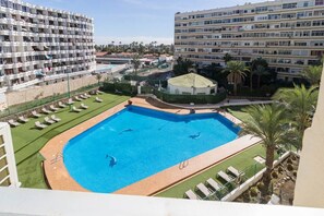 Una piscina con múltiples carriles, rodeada de sillones de descanso y palmeras, con edificios altos al fondo.