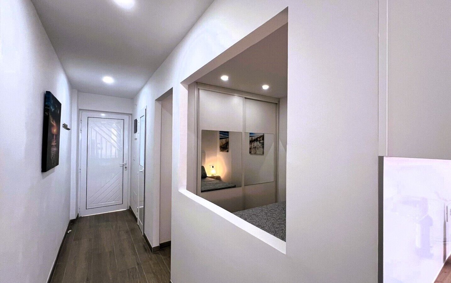 Ein Flur mit einer Tür am Ende, einer kleinen Nische mit einem Bett und einem Nachttisch sowie einem gerahmten Bild an der Wand.