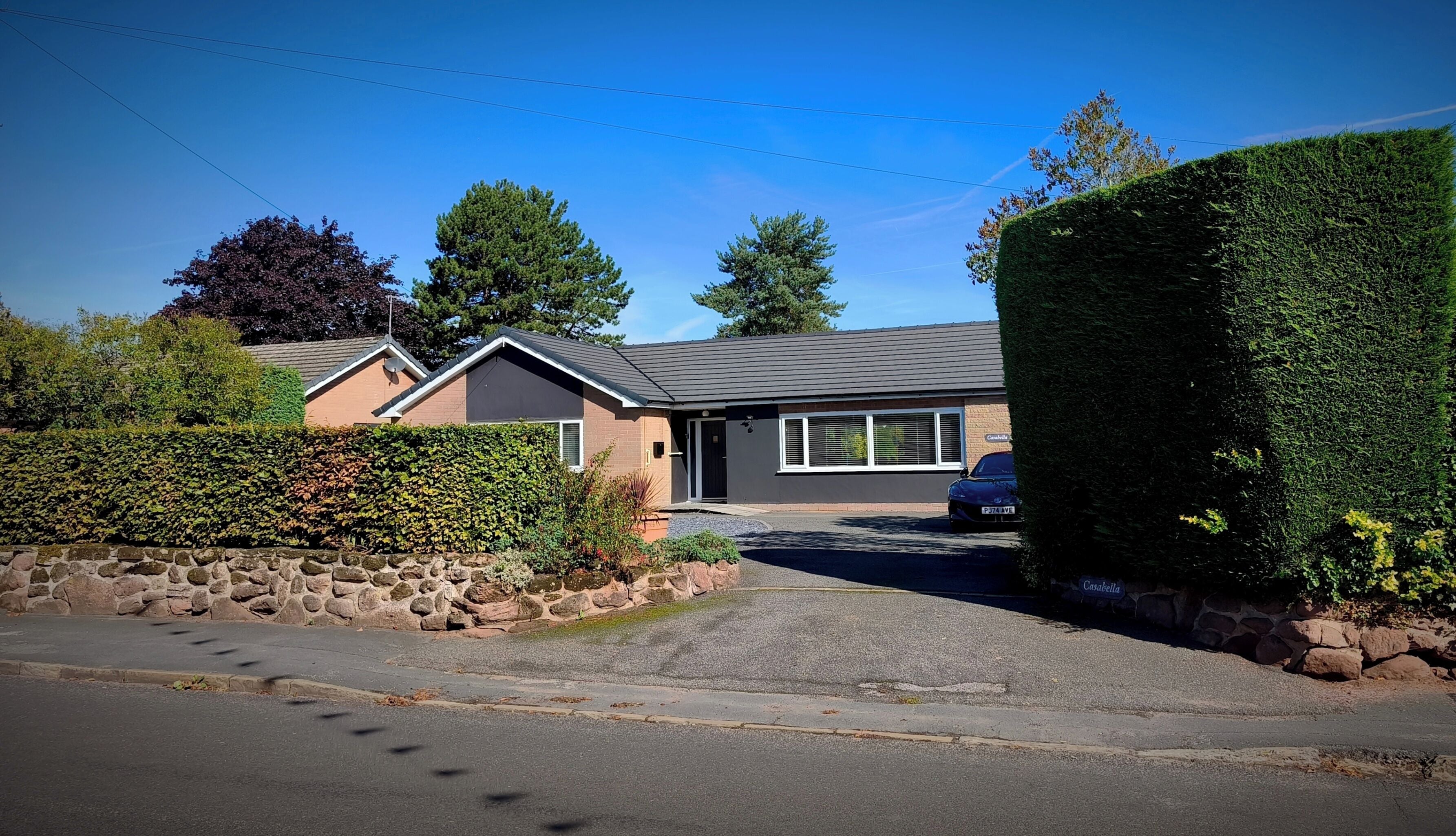 A residential house with a paved driveway, a stone wall, and a well-maintained hedge.
