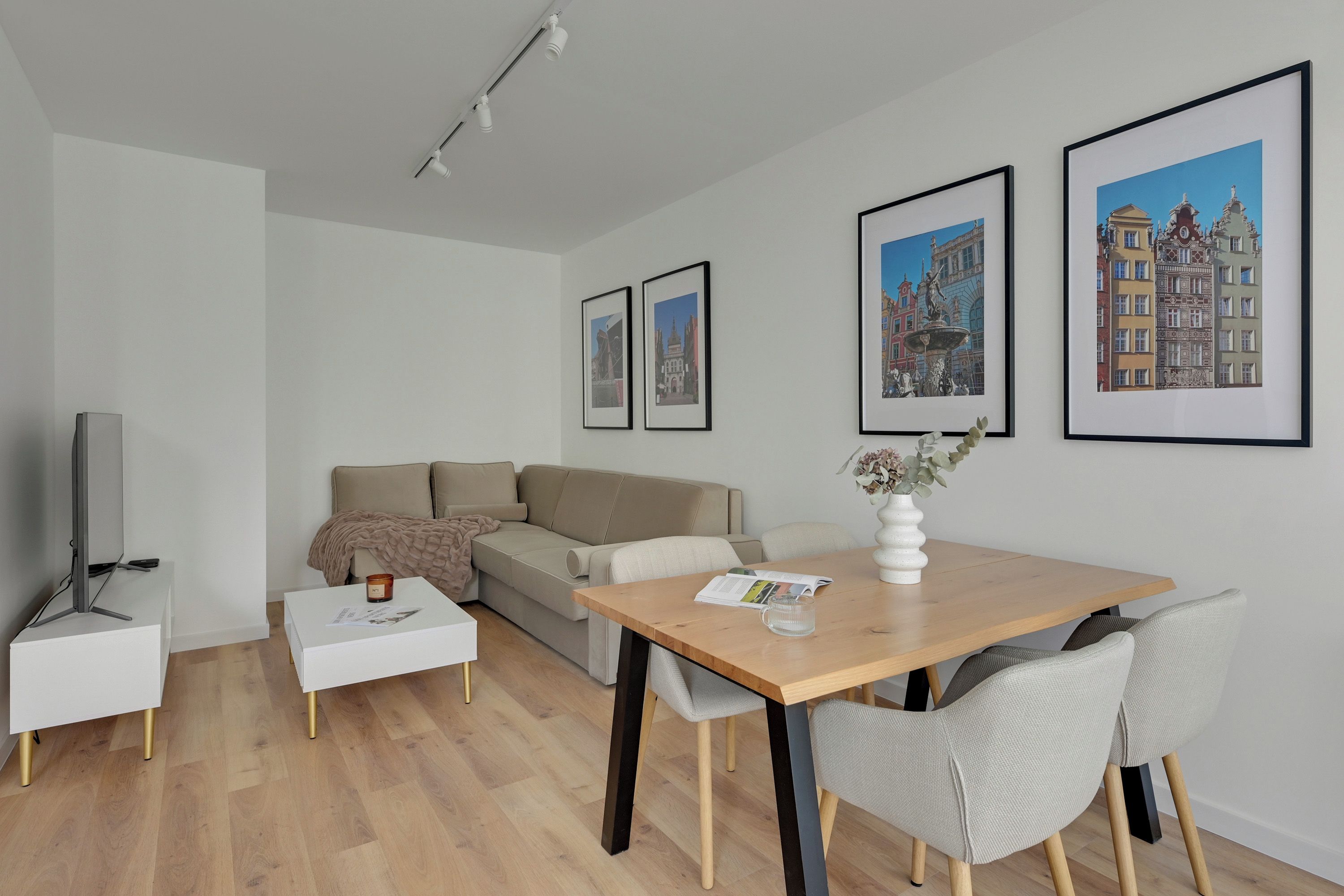 Una sala moderna con sofá, mesa de centro y mesa de comedor con sillas. Hay cuadros en la pared y un televisor sobre un mueble.