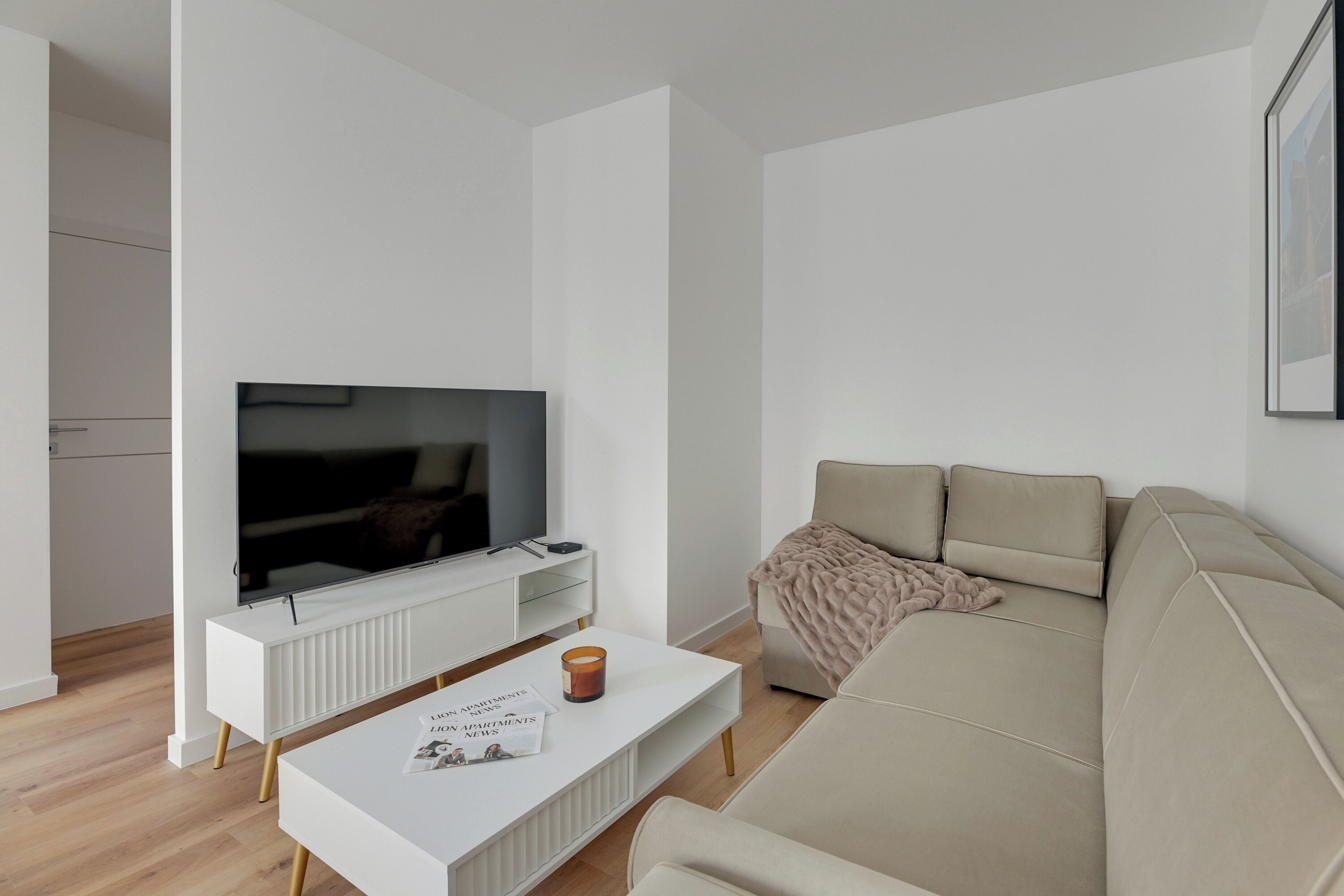 A modern living room with a flat-screen TV on a white stand, a beige sofa, and a white coffee table with a magazine.