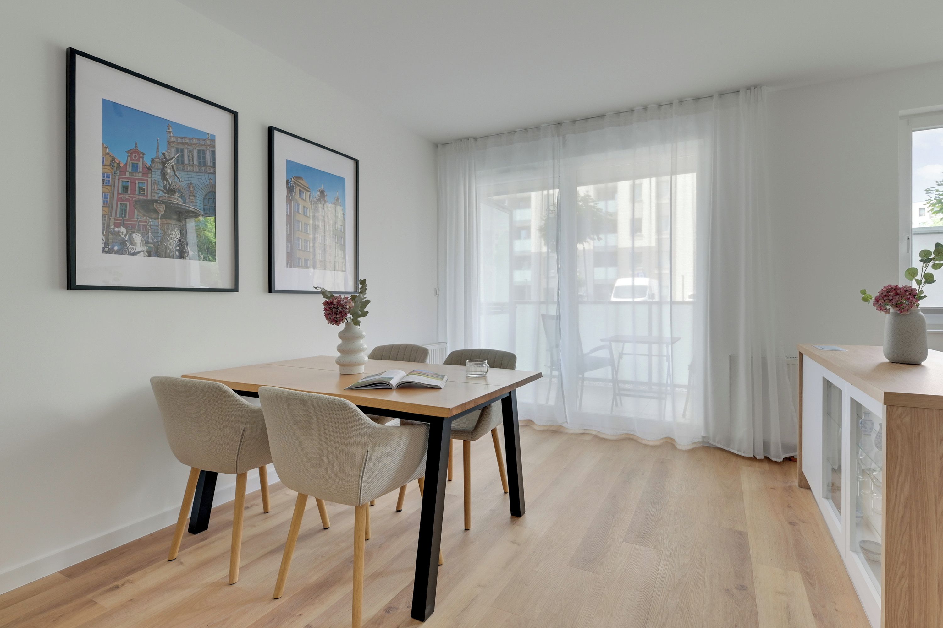 Un coin repas avec une table en bois, quatre chaises, deux tableaux encadrés au mur et une vue à travers des rideaux légers.