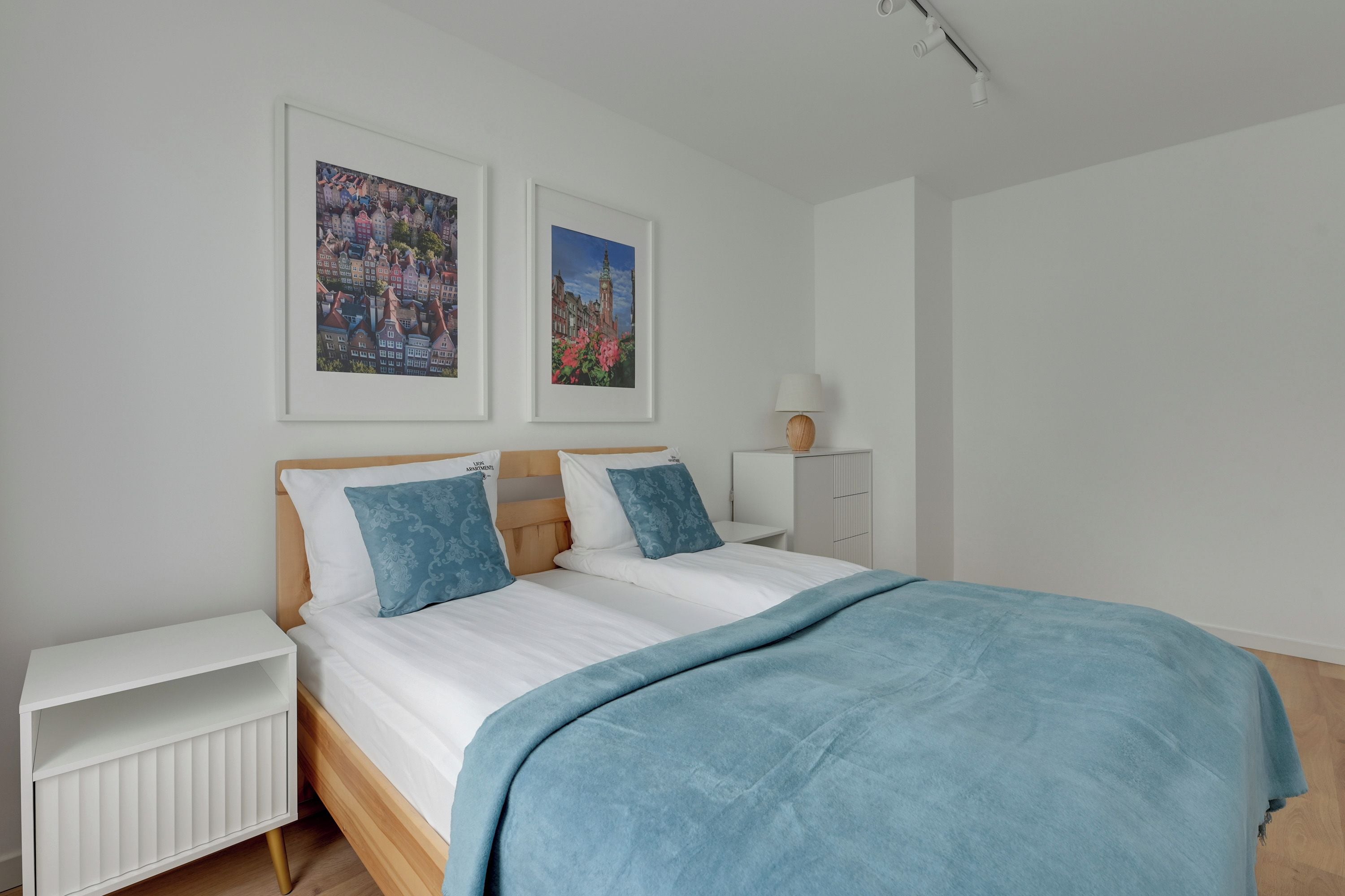 Un dormitorio con una cama, dos almohadas, una mesita de noche, una lámpara y dos cuadros en la pared.
