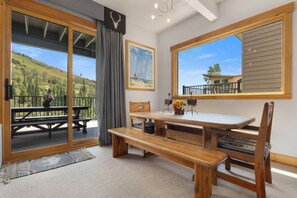 Ein Zimmer mit einem Holztisch, Bänken, einem großen Fenster mit Blick auf ein Gebäude und Bäume sowie einer Tür zu einer Außenterrasse.