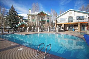 A swimming pool with a wooden deck and metal railings, surrounded by buildings with balconies and a snow-covered tree. - Ski In/Out, Panoramic Views, Pool, Hot Tub, Sauna, Shuttle Service +EZCancel (Steamboat Springs)