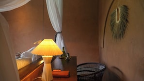 A room with a bed, a bedside table with a lamp, a chair, and a decorative wall hanging.