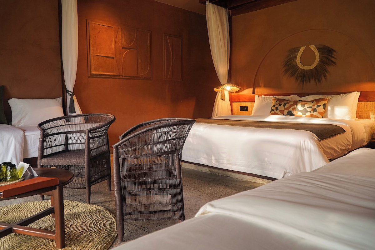 Habitación de hotel con dos camas, una sala de estar con muebles de mimbre y una lámpara fijada a la pared.