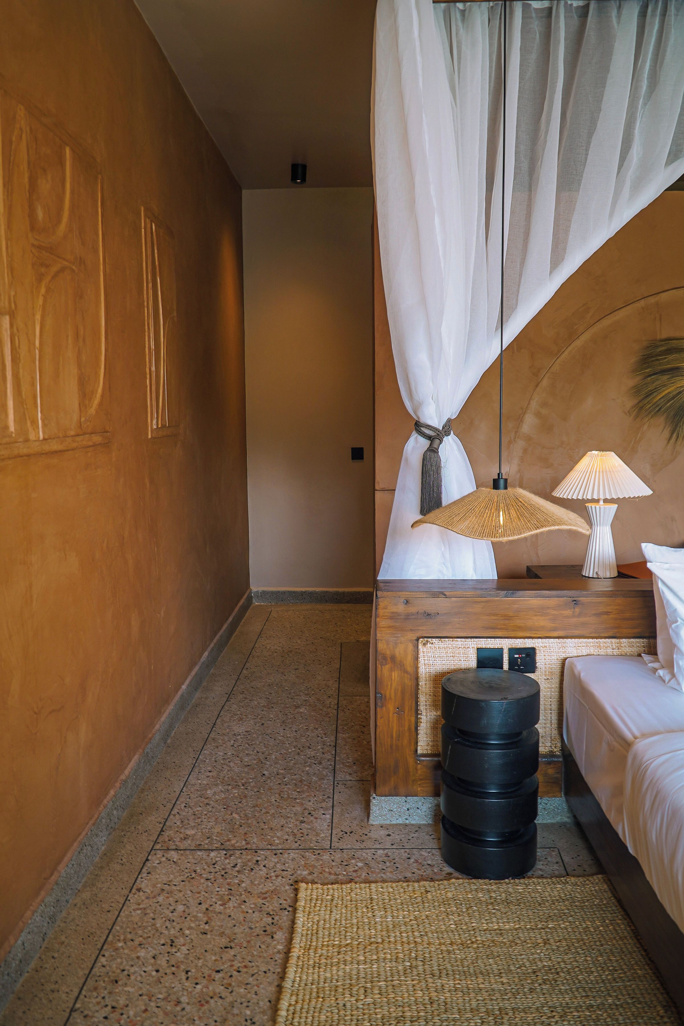 Una habitación de hotel con una cama, una mesita de noche, una lámpara, un tapete tejido y una pared con un panel decorativo.