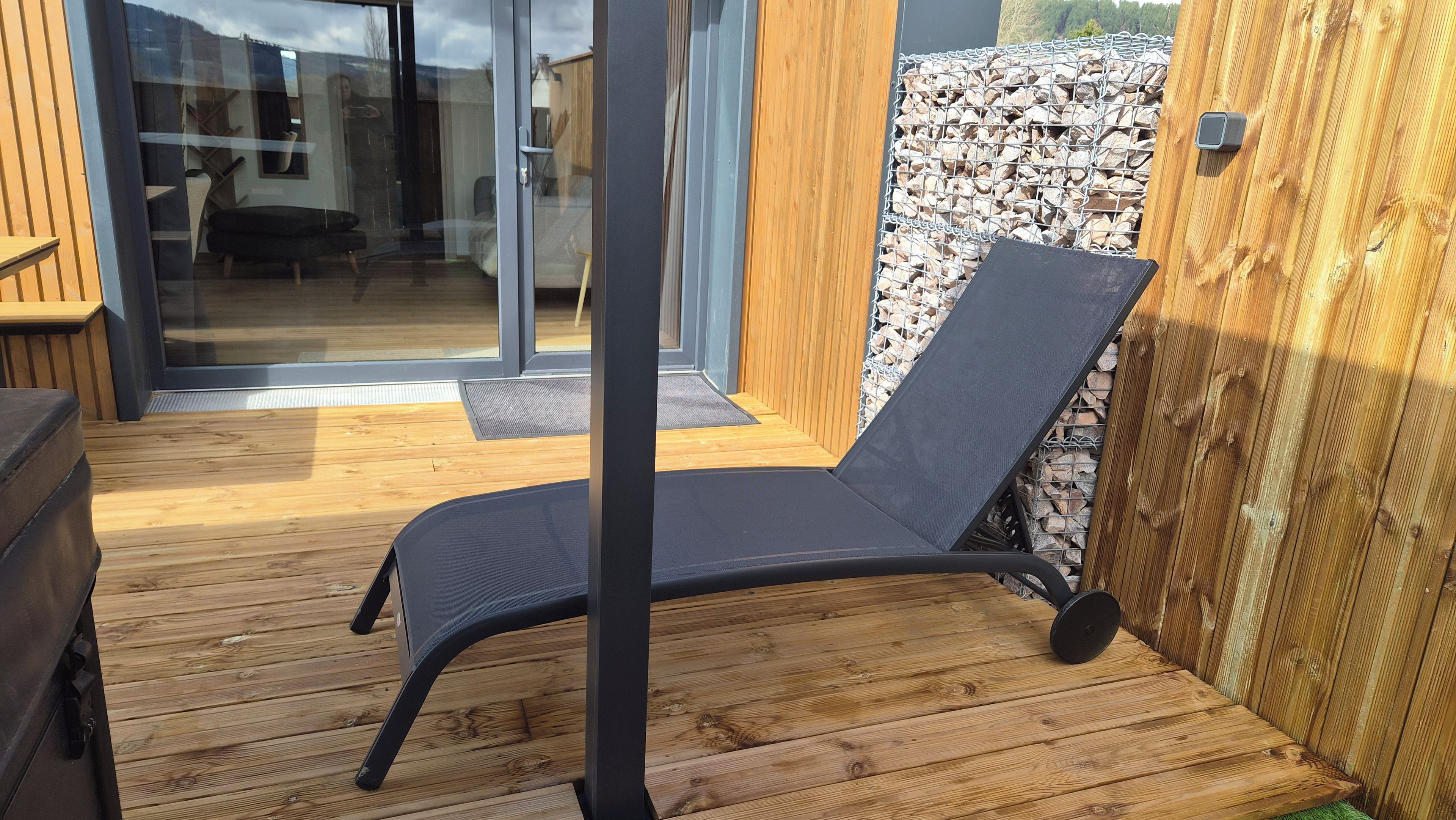 Una terraza de madera con una silla reclinable negra, puertas correderas de vidrio y una pared de piedra.