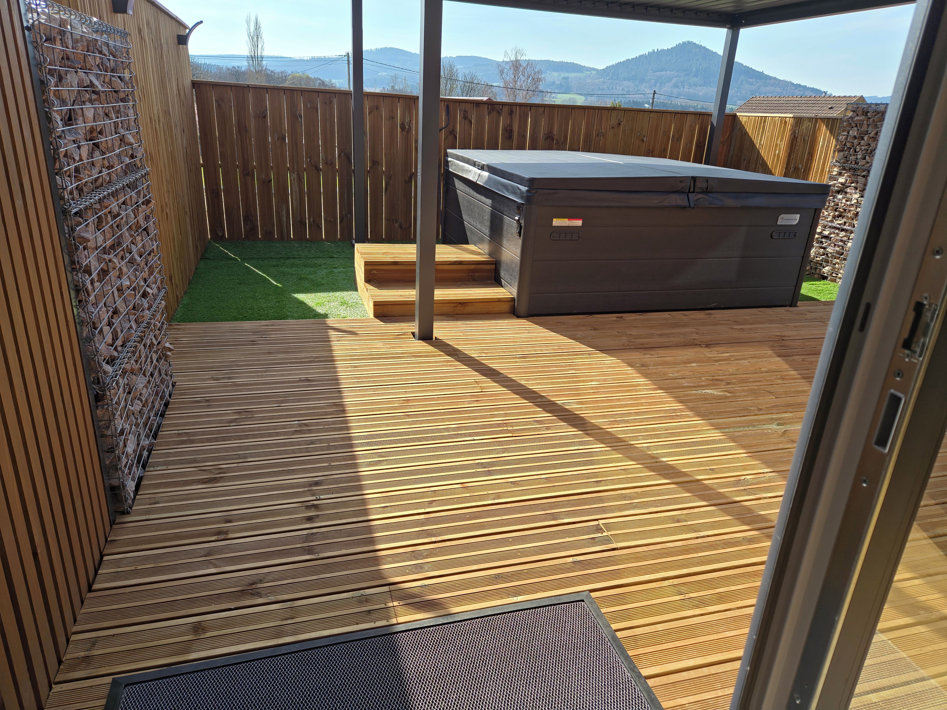 A wooden deck with a hot tub, a small artificial grass area, and a view of mountains.