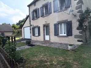 Ein zweistöckiges Haus mit Natursteinfassade, zahlreichen Fenstern mit Fensterläden, einer Garagentür und einem kleinen Garten.