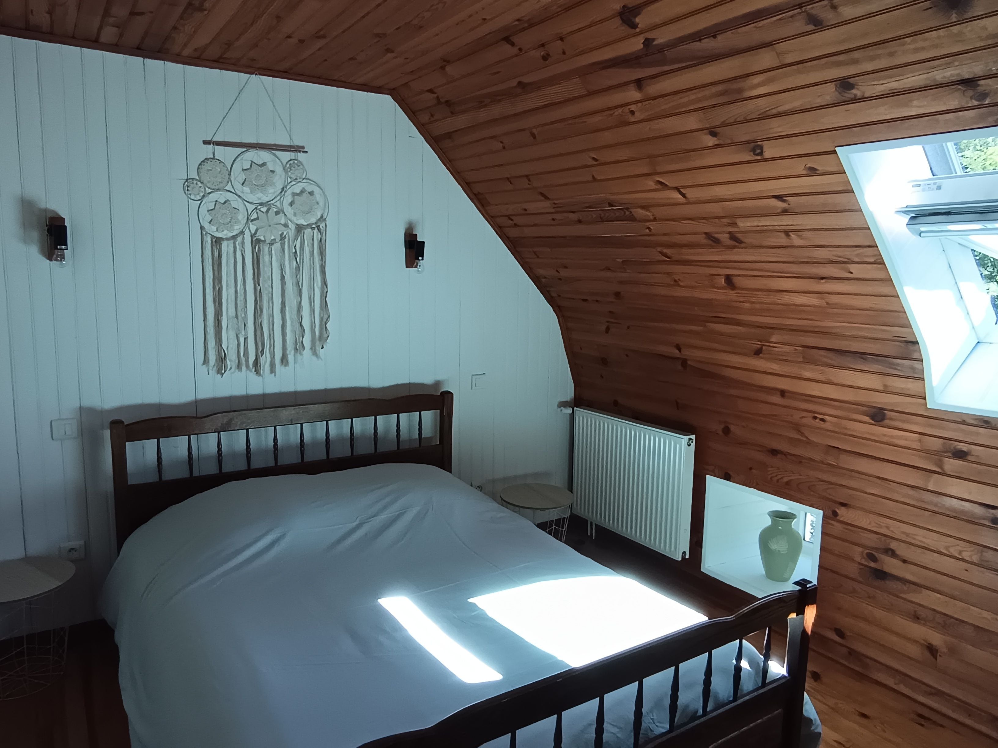 A bedroom with a sloped ceiling, a bed with a wooden headboard, a dreamcatcher, a radiator, and a window with a view.