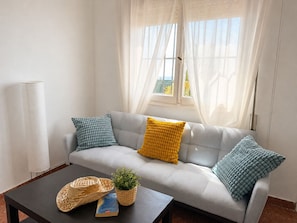 A modern living room with a grey sofa, two patterned cushions, a small table with a plant and a book, and a window with sheer curtains. - Villa Juanita – Andalusian charm near Torrecilla Beach, Nerja (Nerja)