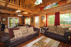 L’intérieur d’un chalet en rondins, avec un escalier en bois, une pancarte de bienvenue, un coin repas et un jeu d’échecs posé sur le sol.