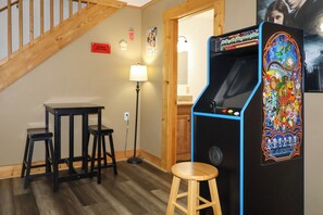 Ein Zimmer mit einer Holztreppe, einem kleinen Tisch mit zwei Hocker, einer Lampe und einem farbenfrohen Spielautomaten.