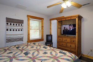 Ein Schlafzimmer mit einem Bett, einem Fernseher, einem Holzschrank, einem Fenster mit Jalousien und einem Deckenventilator.