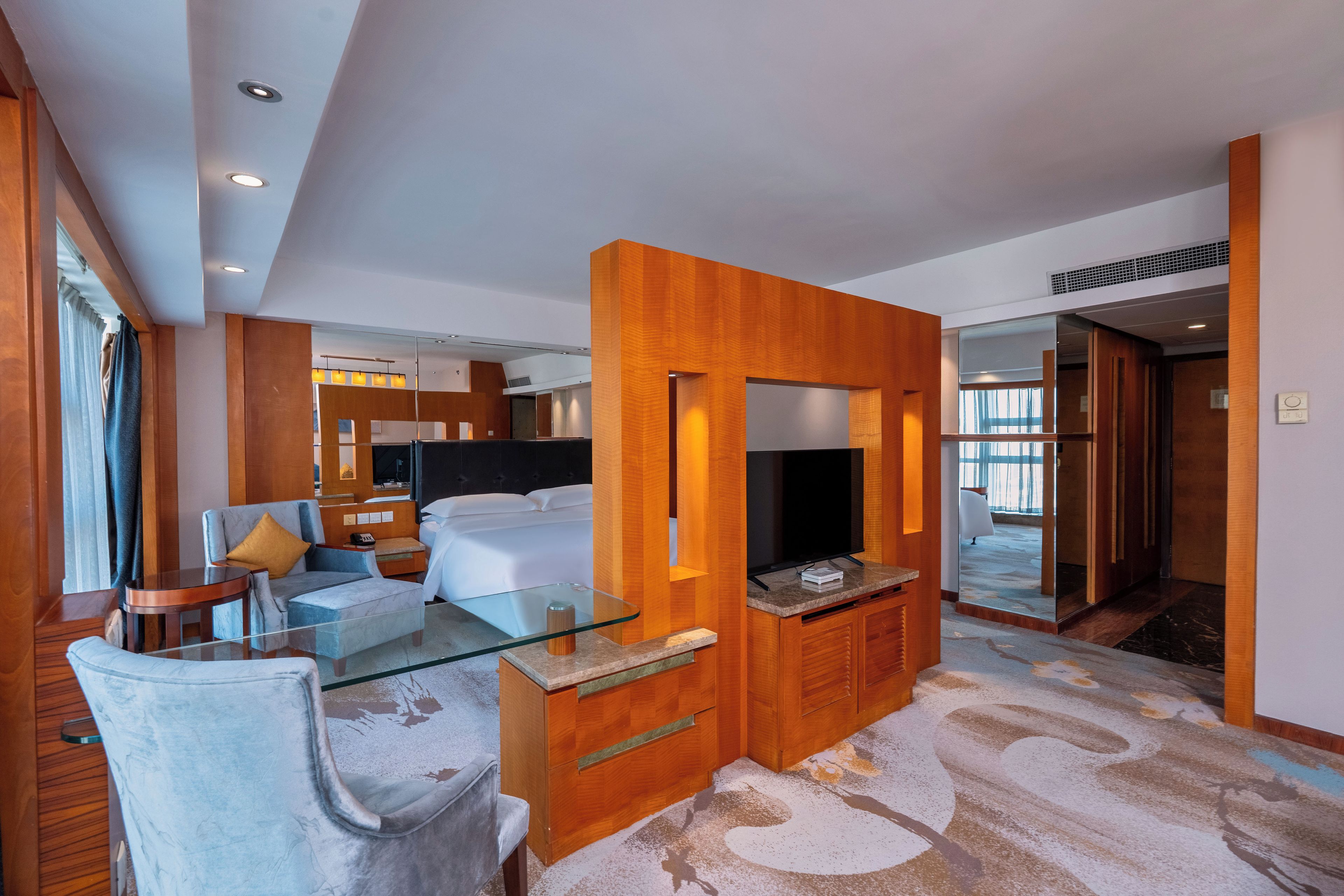 Una habitación de hotel moderna con una mesa de centro de vidrio, un sofá, un televisor y una pared decorativa de madera.