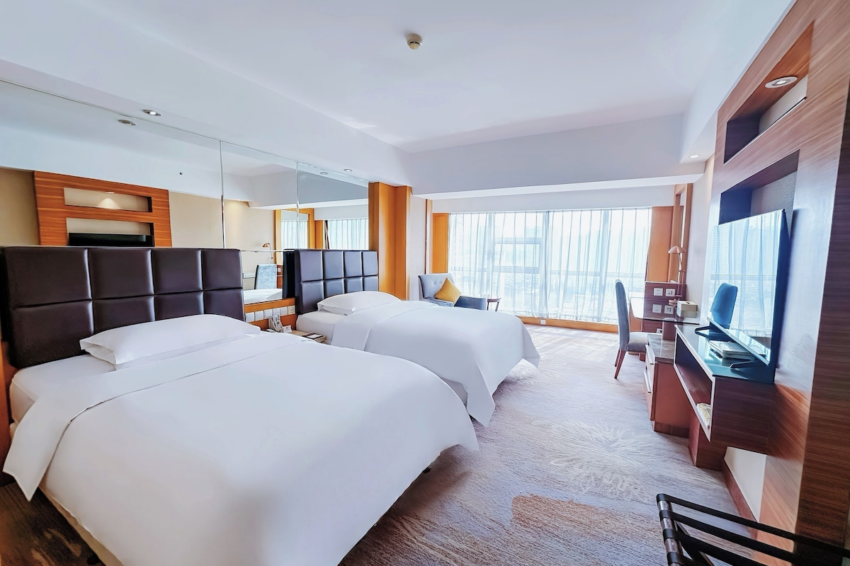 A hotel room with two beds, a desk, a chair, a TV, and a large window.