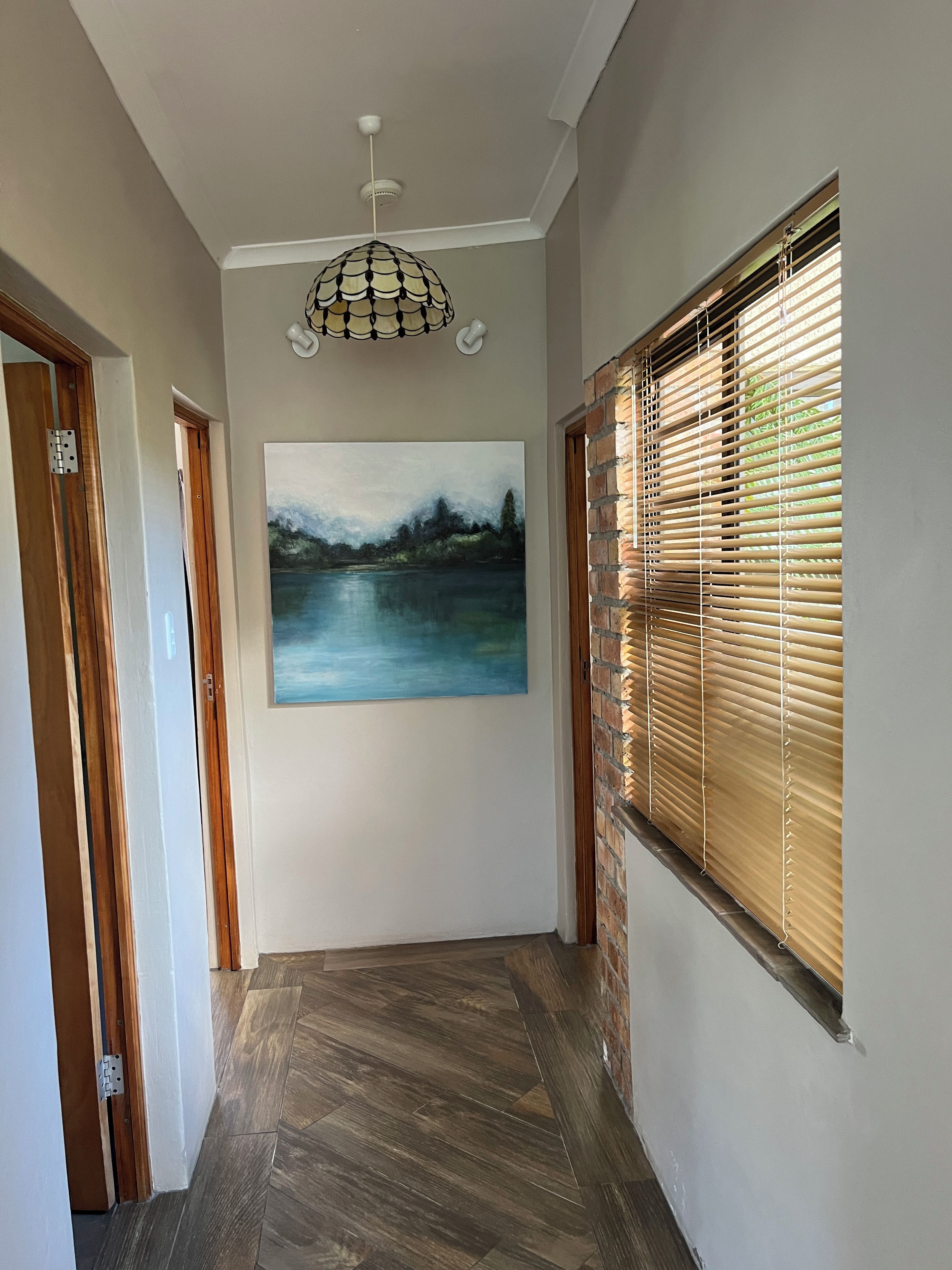 Um corredor com piso de madeira, luminária no teto, pintura de paisagem e janela com persianas.