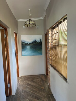 A hallway with a wooden floor, a ceiling light fixture, a painting of a landscape, and a window with blinds.