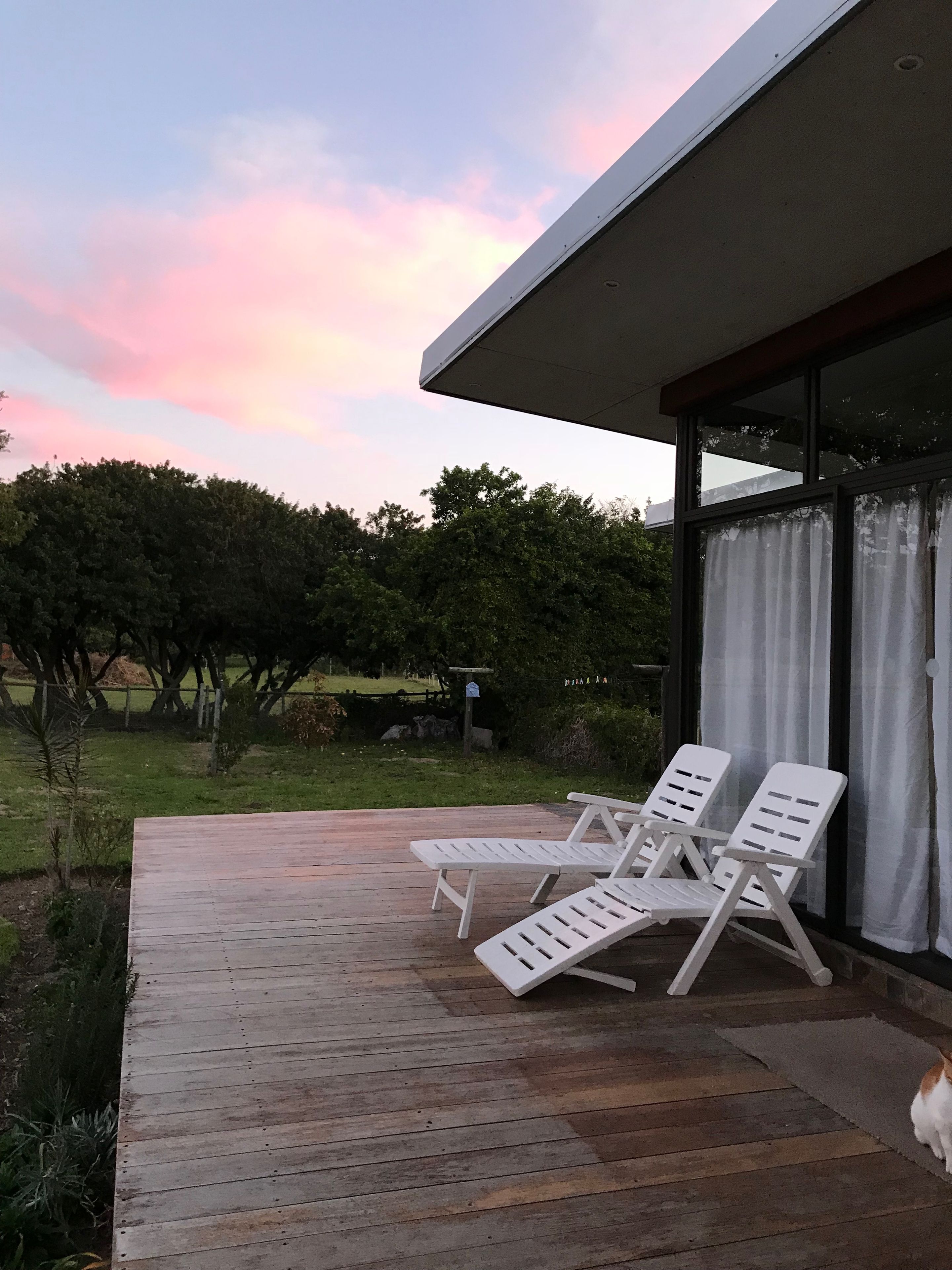 A wooden deck with two white lounge chairs, a glass door, and a view of a grassy area with trees.