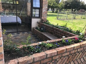 A room with a bed, a window, and a view of a garden with a brick wall and plants.
